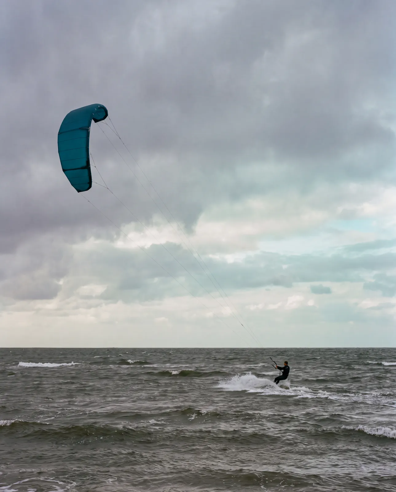 Kite im Aufbau am Wasserrand, Leinen ausgelegt, Schüler von hinten in Neopren, harter Seitenwind, Nordsee-Licht am Vormittag.