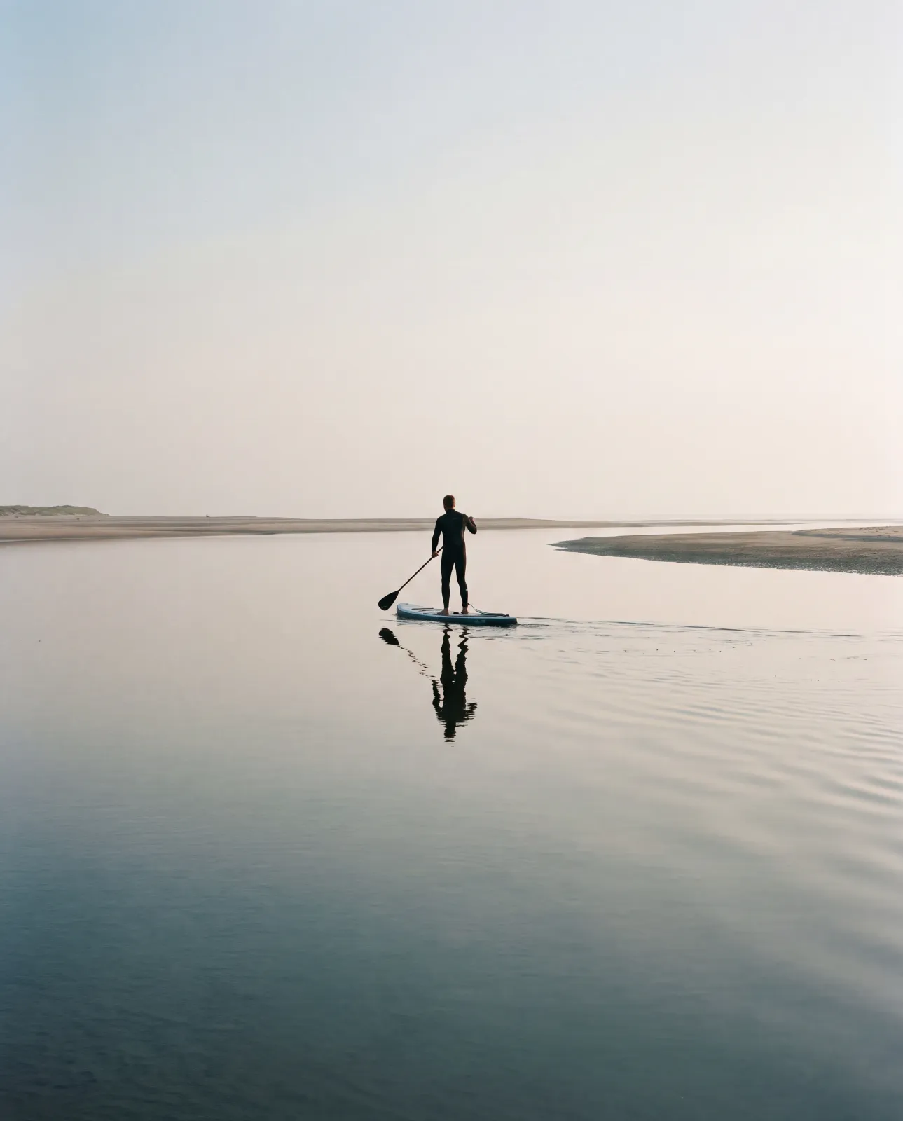 Boards und Paddel am ruhigen Priel bei ablaufend Wasser, spiegelnde Wasserfläche, Person von hinten beim Einsetzen, spätes Licht.