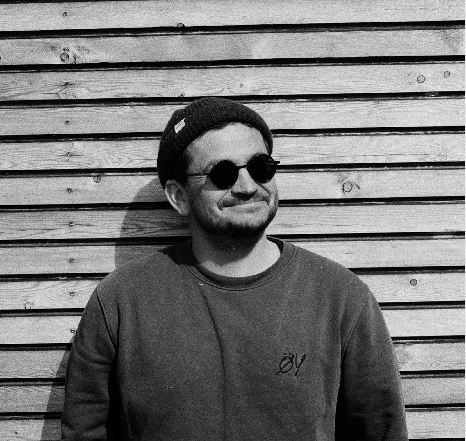Wilke Brinkmann vor der Holzwand der Station, Mütze, Sonnenbrille, Pullover mit ÖY-Logo, schwarz-weiß.